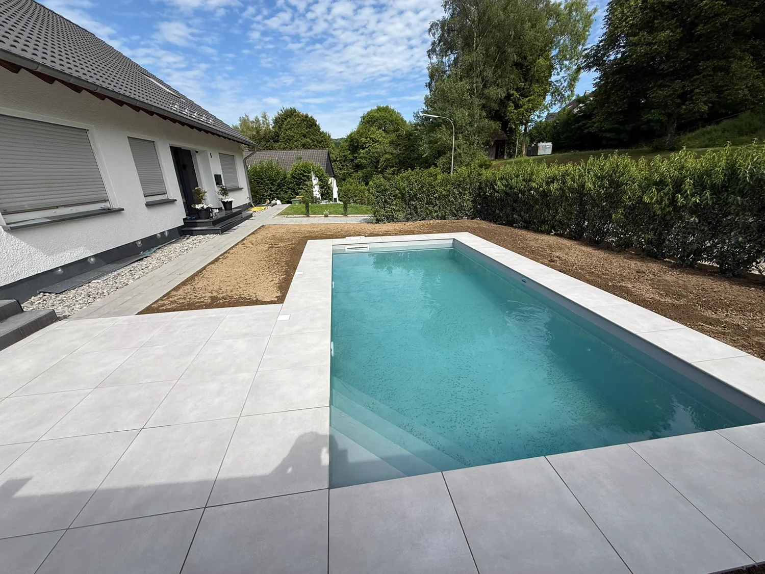 Fertige Poolanlage mit türkisfarbenem Wasser, breiter heller Plattenterrasse und Hecke vor einem Wohnhaus